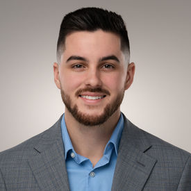 Professional headshot of a man with short hair and beard wearing a gray blazer, smiling against a neutral background