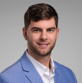 Professional headshot of a man with short hair wearing a light blue blazer, smiling against a neutral background