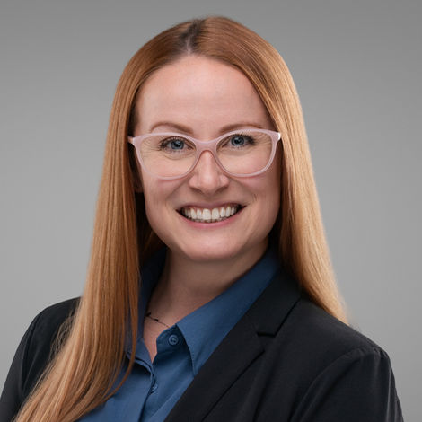 Professional headshot of a smiling woman with long red hair and glasses wearing a blazer, gray background.
