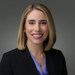 Professional headshot of a woman with blonde hair wearing a black blazer and purple top, smiling against a gray background