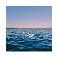 Astronaut floating on calm ocean at sunset with a pastel sky. The mood is peaceful with deep blue water and distant shoreline.