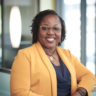 Dr. Shelitha Williams from SUNY Genesee Community College. Smiling and wearing an orange blazer adorned with a gold medallion necklace.