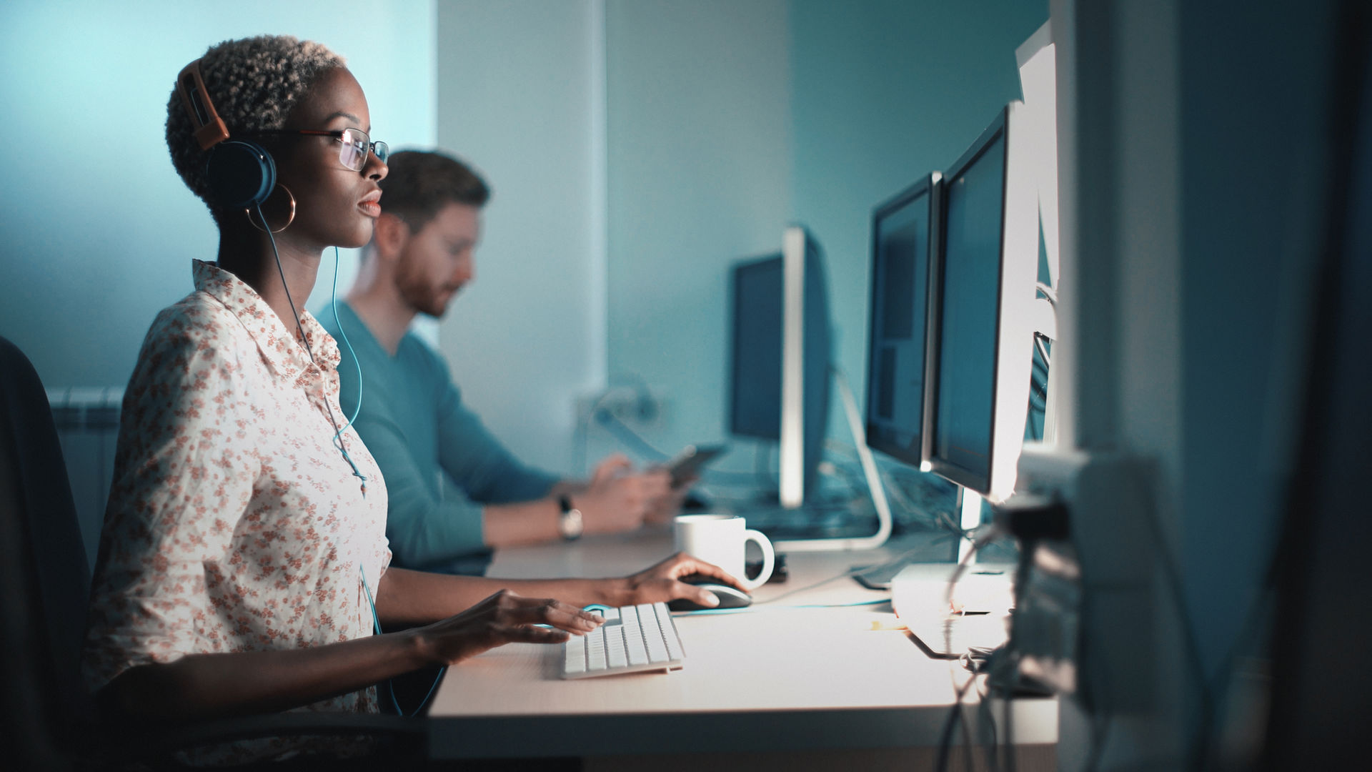 A corporate style photography of focused female professional wearing headphones and glasses, working on a computer in an office setting. In the background, a male colleague is also engaged with his computer and phone. The image highlights a modern, collaborative work environment, showcasing concentration and productivity of corporate culture.
