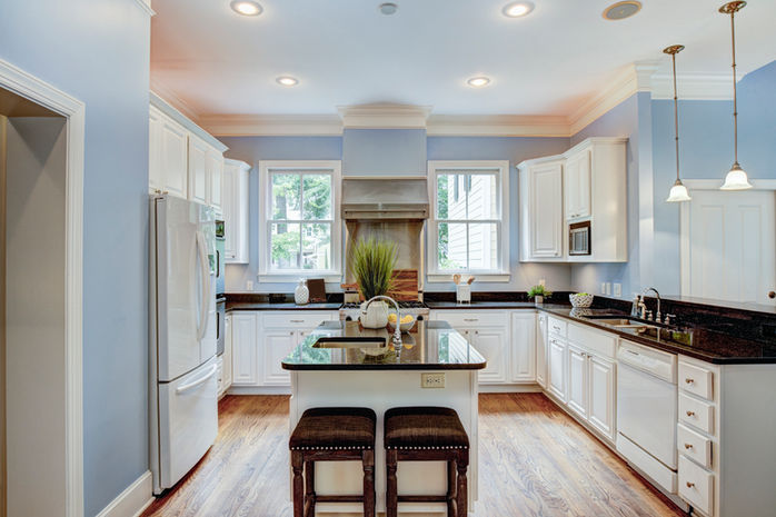 A classic elegant kitchen complimented by light blue walls, traditional white cabinets and glossy shiny stylish granite countertops. Hanging lights and white trimmed windows illuminate the room decorated with various kitchen decorations and modern stainless steal appliances.