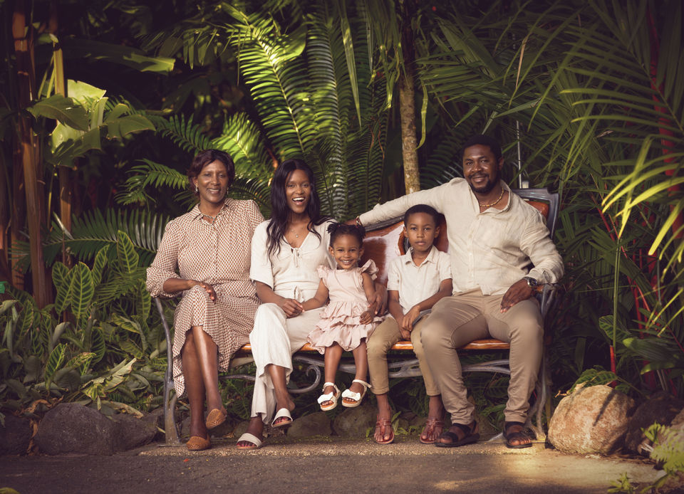 A three-generation family sits closely together on a bench in the Cairns Botanic Gardens, with verdant tropical plants in the background. They smile serenely, exuding a sense of familial warmth and contentment.