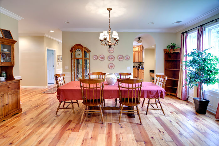 A traditional dining room with antique wooden features including polished wooden shelves displaying unique cutlery and plates. A red table cloth covers a wooden dining table with a traditional smaller chandelier hanging over it. Arched entrances feed into the brightly lit room filled with natural light and accompanied by a timeless wooden floor.