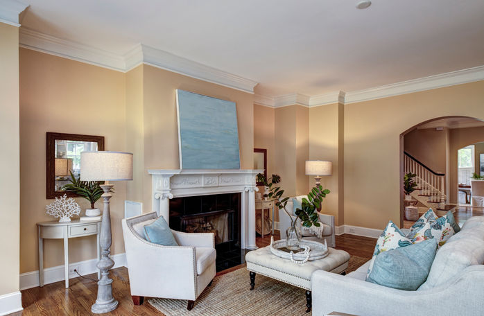 A traditional elegant decorated living room with a classic fireplace surrounded by stylish beige off-white furniture with light blue accents. The luxury living room is decorated with unique lights and art work placed across the brightly lit room with beige walls.