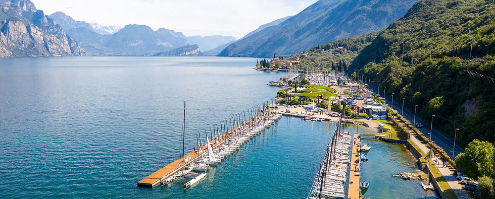 A marina on a serene lake with rows of docked sailboats, surrounded by mountains and greenery under a clear blue sky.