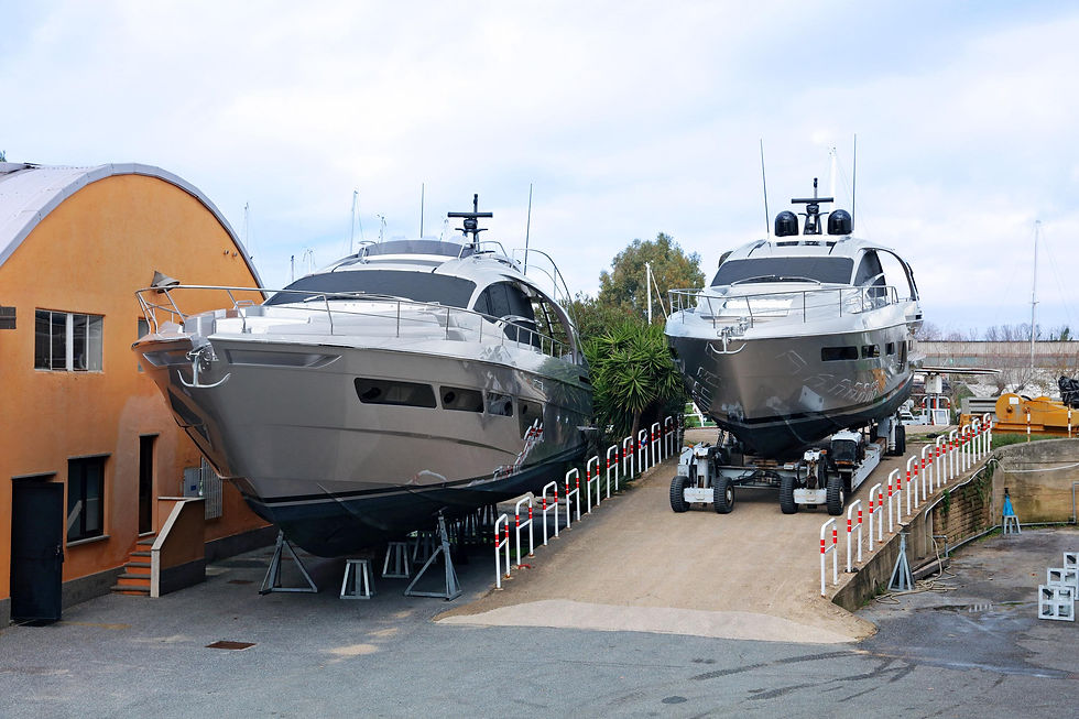 Two large gray yachts on stands in a shipyard. An orange building is on the left, with trees in the background and a cloudy sky.