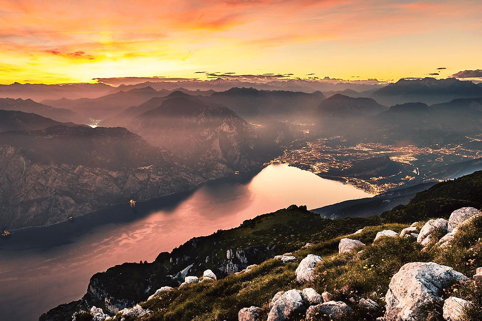 Sunset over mountains and lake, with warm orange and pink hues illuminating the sky. City lights twinkle below; foreground rocks and grass.