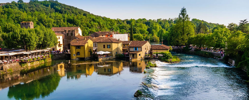 Colorful riverside village with yellow houses, cafes, and people by a small waterfall, backed by lush green hills.