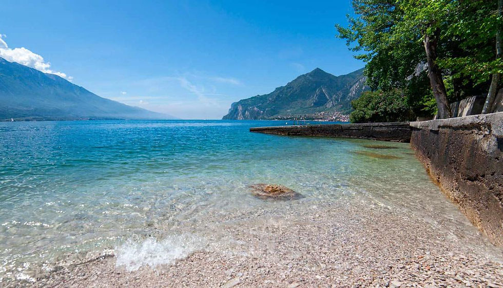 Clear blue lake with pebbly shore, stone seawall, and mountains under a sunny sky; calm, serene scene.