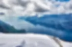 Snowy mountain overlook above a blue lake and rugged peaks under dramatic clouds, with a wooden fence in the foreground.