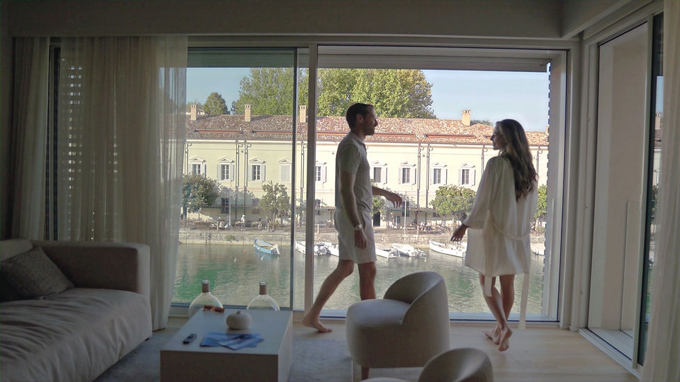 A couple in white loungewear stands by large windows overlooking a canal and building, with a cozy interior setting and soft lighting.