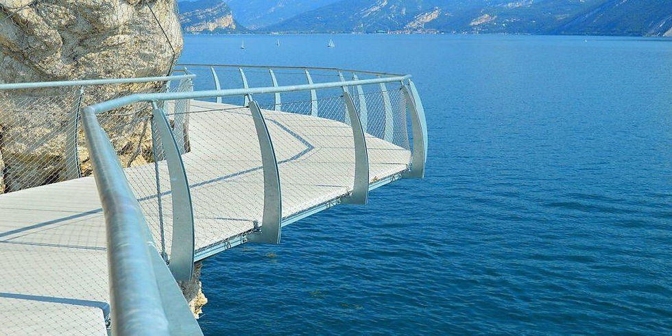 Curved steel walkway over blue lake, lined with railings; rocky cliffside and distant mountains in background. Peaceful and scenic view.
