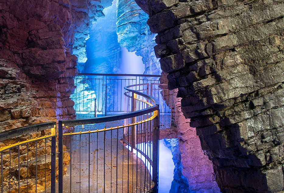 Spiral walkway in a cave with colorful lighting. Blue and purple hues illuminate rocky walls, creating a mystical atmosphere.