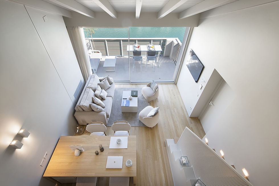 Modern living room with beige furniture, wooden floor, and balcony view. Dining table set with laptop and cups. Bright, airy atmosphere.
