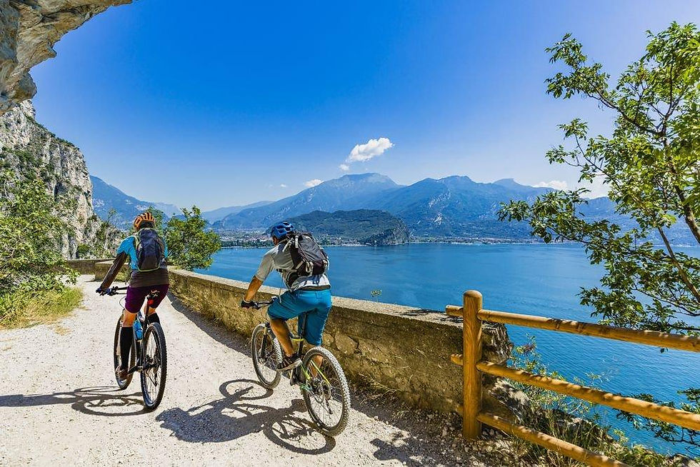 Two cyclists ride on a scenic path overlooking a blue lake and mountains on a sunny day, surrounded by greenery and rocky cliffs.