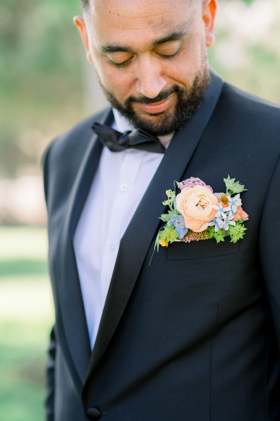 Portrait of a groom in a dark suit looking down at a peach and pastel boutonniere on his lapel