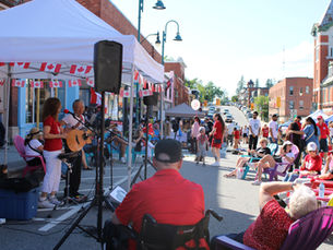 Crowd enjoying a downtown Canada Day street festival with tents, live music, Canadian flags, and historic buildings in the background.