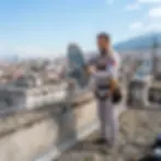 Outdoor photography of a technician installing a satellite dish on a city rooftop. He is wearing a safety harness with urban buildings in the background.