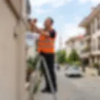 Outdoor photography of a technician on a ladder installing a security camera on a building's exterior wall. Professional service in a residential street.