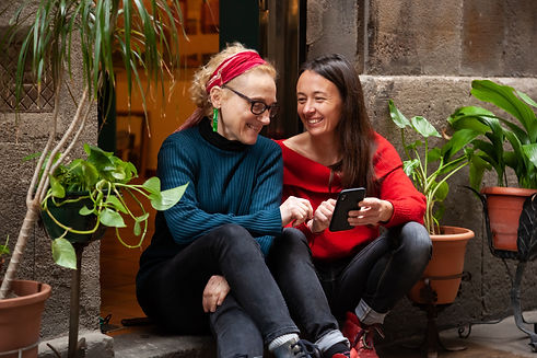 A joyful moment between friends and neighbors sitting outside, smiling and laughing while looking at a smartphone. Surrounded by potted plants and warm, rustic stone walls, this heartwarming image captures genuine connection and shared happiness in a community of oneness.