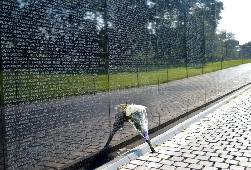 Light reflects off the vietnam war memorial showing inlays of soldiers name who have passed, with flowers resting against the public art installation.