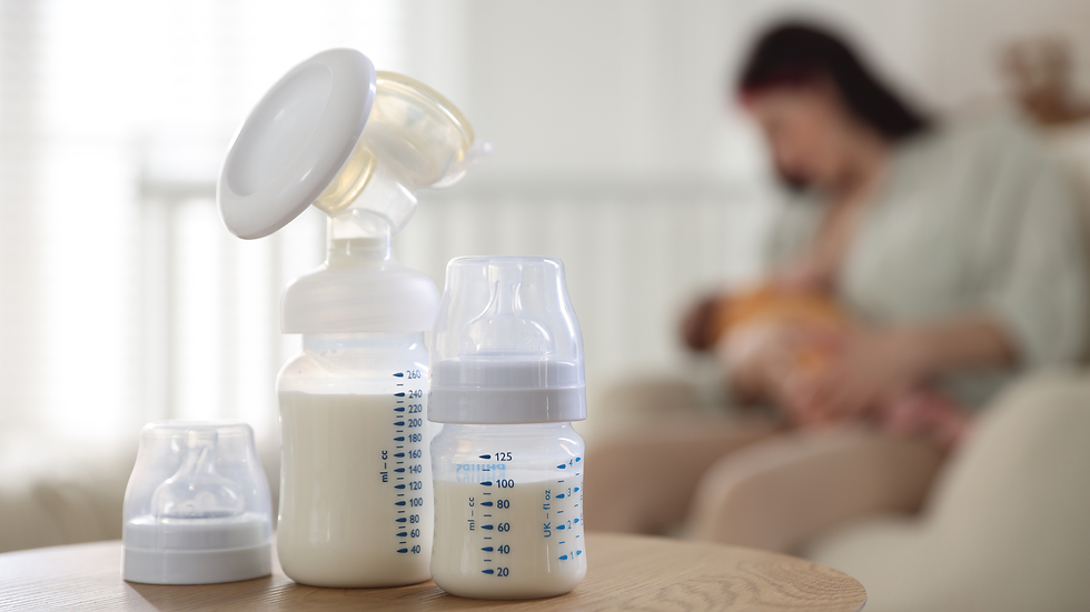 Breast pump and bottles on table; blurred background shows a woman breastfeeding a baby in a softly lit room.