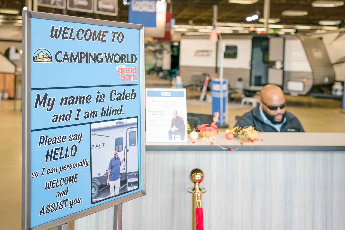 A man with visual impairment at his job. A sign in front of his desk reads: Welcome to Camping World. My name is Caleb and I am blind. Please say hello so I can personally welcome and assist you. End of text on sign.