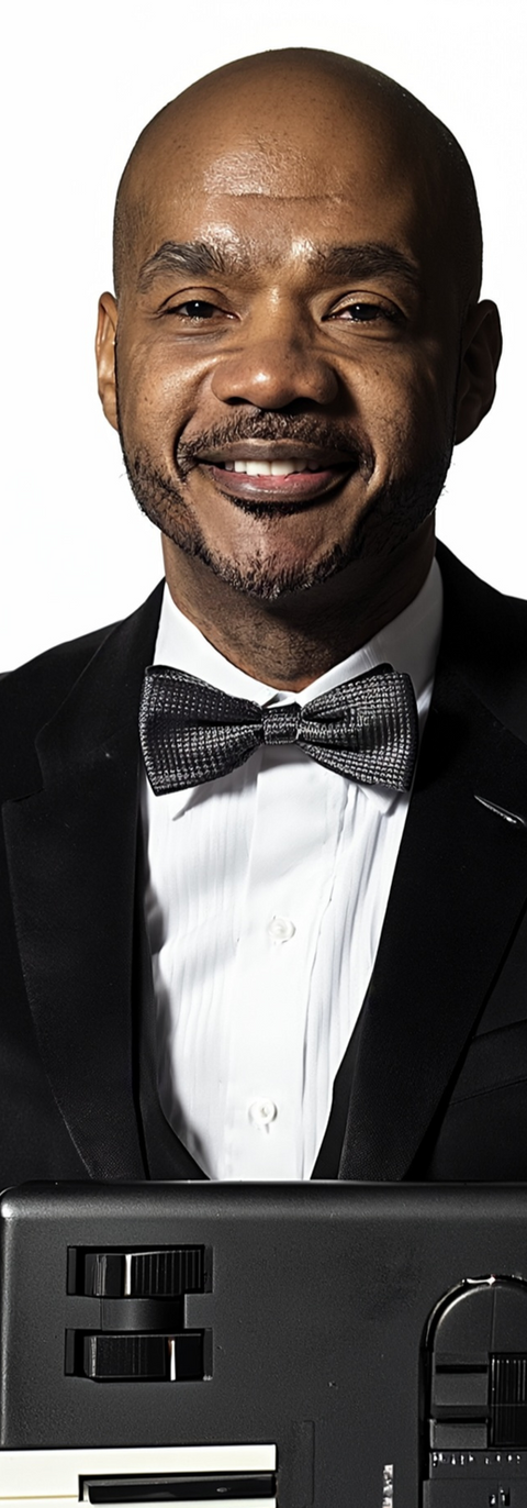 Headshot of Clarence “CC” Johnson, an African American man wearing a black tuxedo and bow tie, smiling while holding a keyboard.