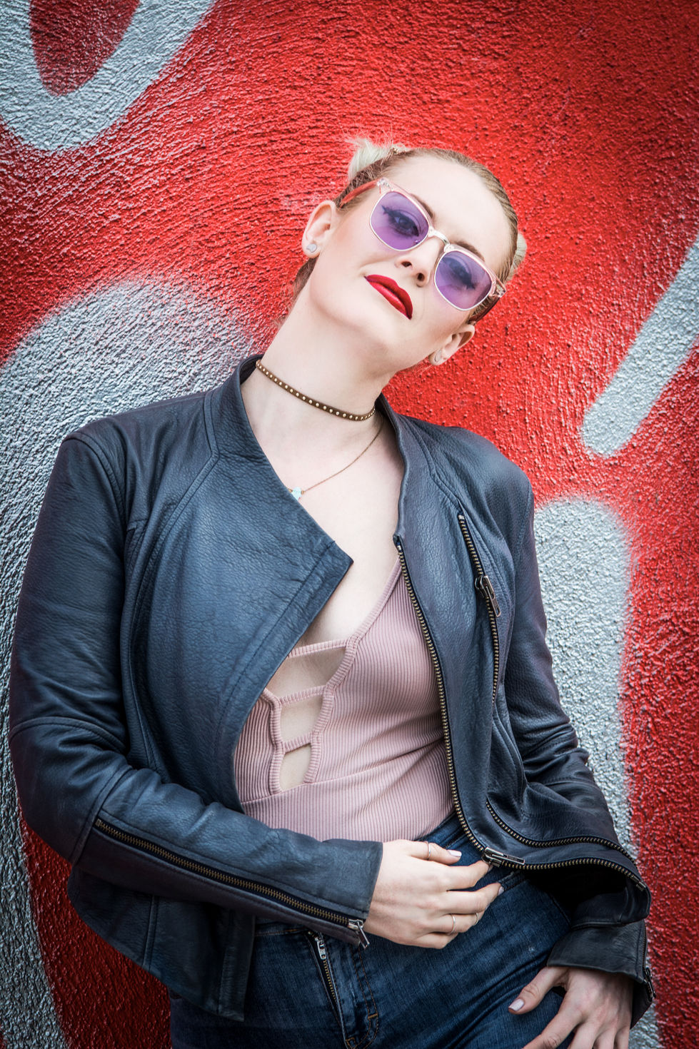 a woman wearing sunglasses and a choker stands in front of a red wall