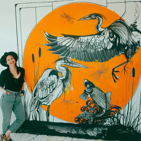 Mural featuring cranes and a fish with the artist Joni Belaruski standing in front