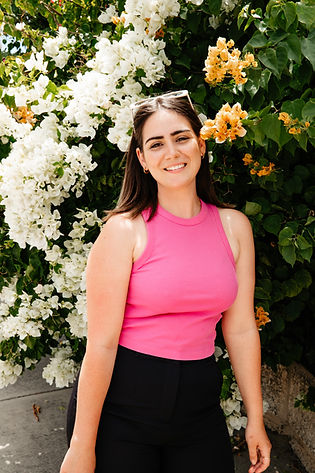 Young women smiling in pink singlet standing in front of flowering bush. Flowers on bush are white and orange.