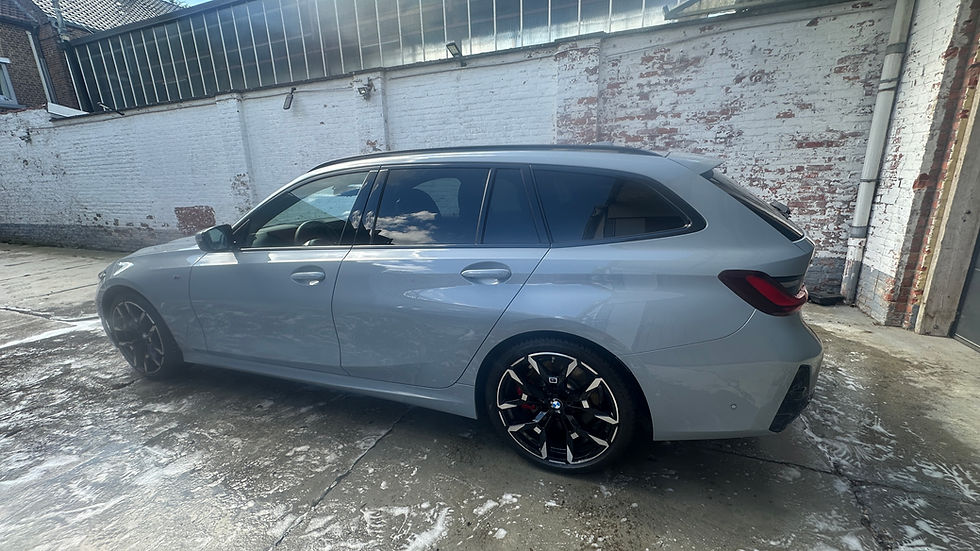 Lichtgrijze BMW 330i M Touring Brooklyn Grau Metallic in zijaanzicht, toont elegante Touring-lijnen en M Sport design, rode remklauwers en sporty velgen, witte bakstenen gebouw op achtergrond