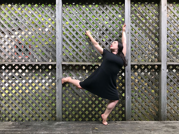 Marissa Finkelstein balances in forced arch against a wooden lattice privacy fence. She wears a black dress and looks up and back to the top of the fence.