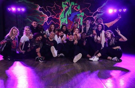 A group of dancers pose in a dance studio in front of a graffiti mural.