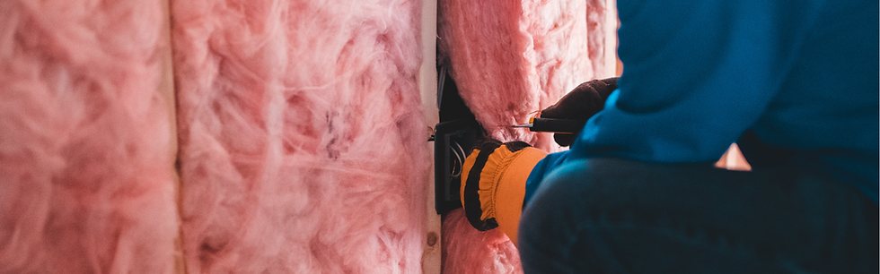 Person in blue and orange installing pink insulation. Close-up of hand pressing material into wall. Dimly lit indoor setting.