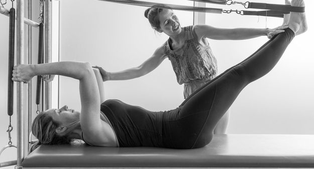 Instructor helps woman perform Pilates exercise on a reformer machine.