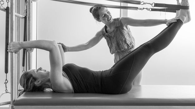 Instructor helps woman perform Pilates exercise on a reformer machine.