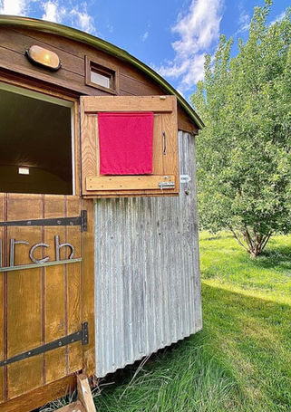Wooden shepherd's hut with 'BII ch' door, red curtain, green landscape.