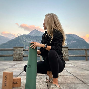 Julia hockt vor einer Bergkette, hält ihre Yoga Matte in der Hand und schaut nachdenklich in die Ferne.