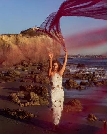 Model on a beach with a flowing purple fabric and rocky formations.