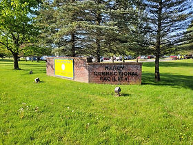 Brick sign reading Marcy Correctional Facility beside a State of New York sign panel, on a lawn among trees