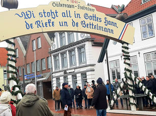 Mehrere Personen stehen auf dem Platz vor dem Alten Pastorat in Heide und beobachten das feierliche Aufstellen des mit Tannengrün geschmückten Eggensymbols der Süderegge. Darüber ein großer Spruchbogen mit plattdeutscher Inschrift.