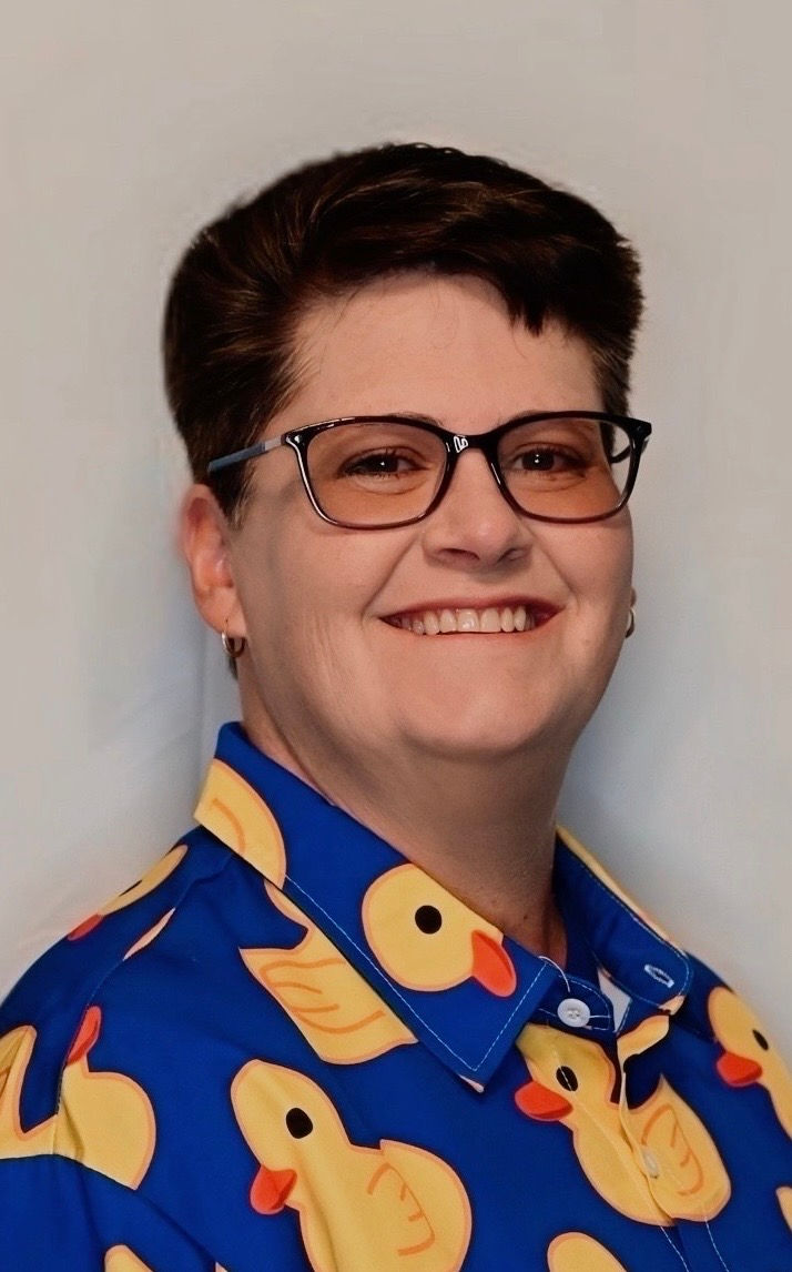 Portrait photo of a smiling adult wearing glasses and a blue shirt with yellow rubber ducks