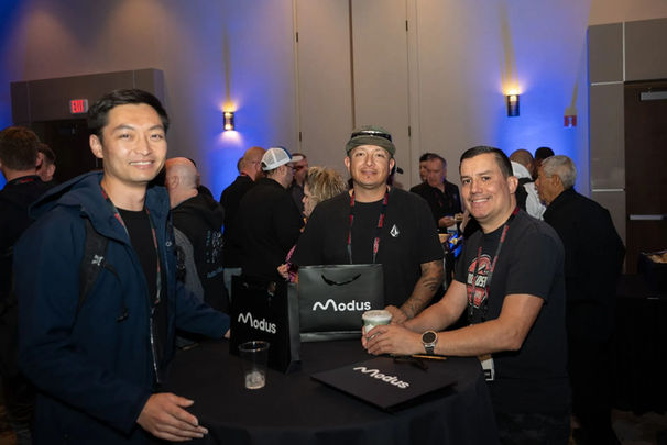 Three men smiling at Modus event with logo and black bags.