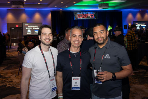 Three men smiling, wearing lanyards, at an event with MASTERTECH EXPO signage.