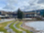 Mountain lodge with large trees, stone paths, and a partially frozen pond in the foreground. Snowy peaks under a cloudy sky set the scene.