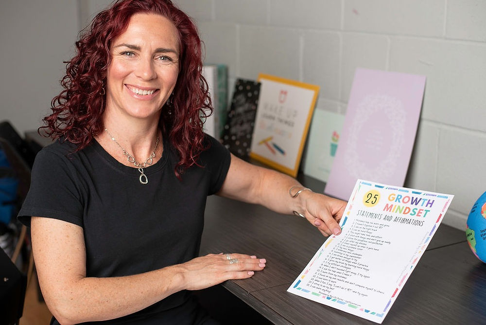 Emmy ( a White woman with curly red hair) is sitting at her desk, looking at the camera and smiling, holding a page of affirmations in her hand.